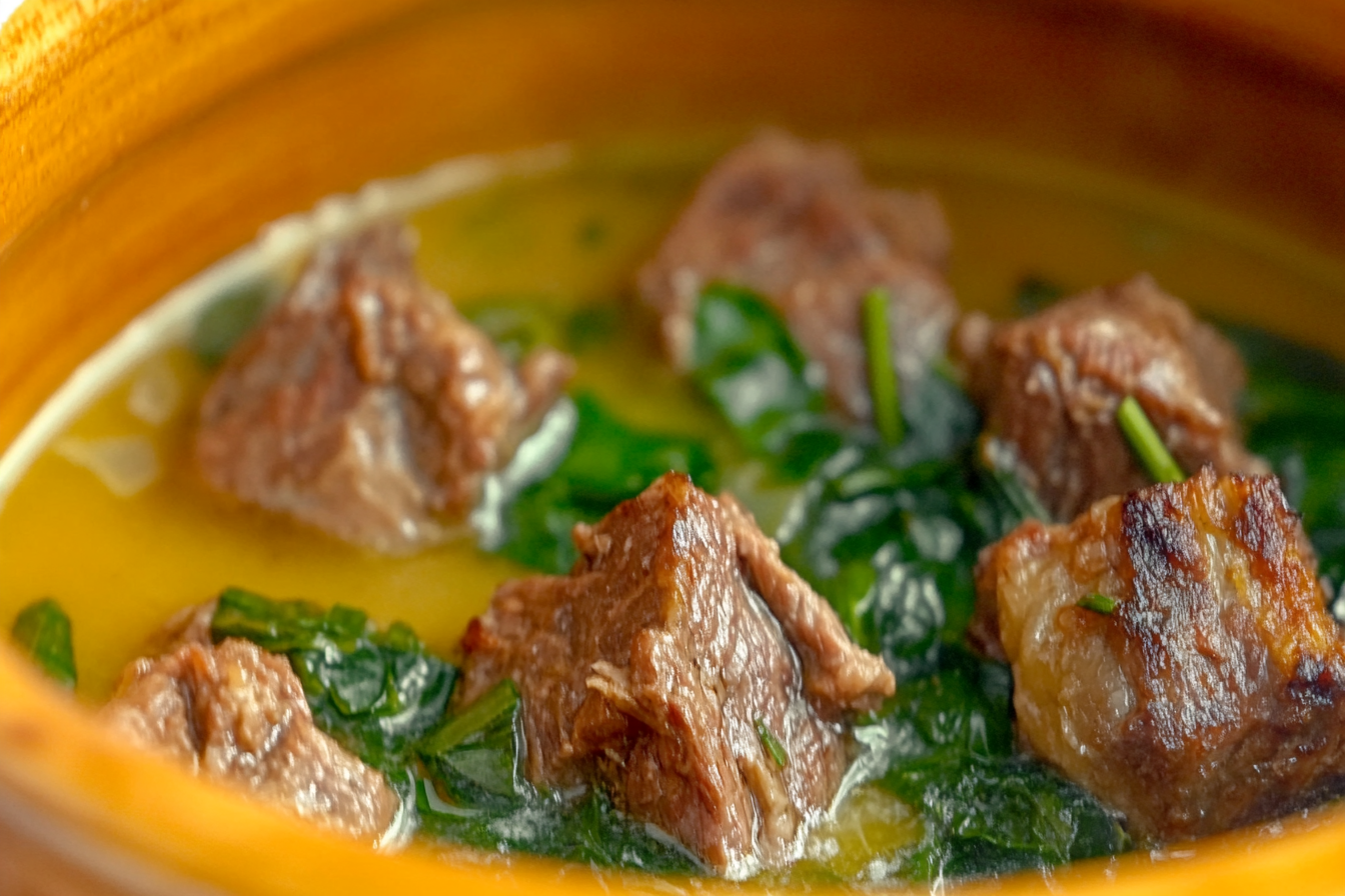 Yakın planda Şevket-i Bostan yemeği; zeytinyağında pişmiş kuzu eti ve yabani otlar, hafif limon aromasıyla servis edilmiş. Yeşil ve altın kahverengi tonlarında, otların ve etin dokusu net şekilde görülüyor. Rustik bir seramik kasede, doğal ışıkla aydınlanmış.